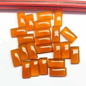 Puede incluir: Una colección de gemas rectangulares pulidas de color naranja. Las piedras están dispuestas sobre una superficie blanca, con un lápiz rojo visible en el fondo. Las gemas tienen una calidad translúcida, con un acabado brillante.