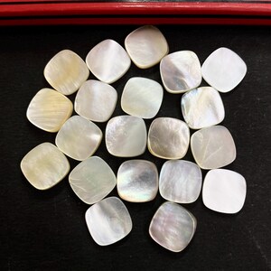 May include: A collection of square, iridescent mother-of-pearl beads. The beads have rounded corners and vary in colour from white to pale yellow, with a shimmering, pearlescent effect. They are arranged on a dark surface.