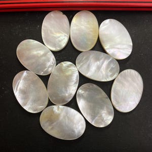 May include: A collection of eleven oval-shaped, iridescent mother-of-pearl cabochons. The polished surfaces reflect light, displaying a range of pearly white and subtle color variations. These gemstones are arranged on a dark surface.