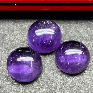 Könnte beinhalten: Drei polierte, runde Amethyst-Edelsteine. Die Edelsteine haben eine tiefviolette Farbe mit glänzendem Finish. Die Steine werden auf einer grauen Oberfläche präsentiert. Das Bild ist gut beleuchtet und hebt die Klarheit und Farbe der Steine hervor.
