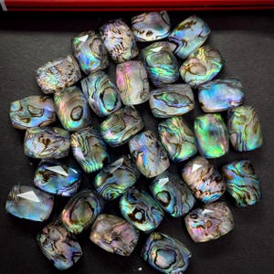 May include: A collection of iridescent abalone shell beads. The beads are rectangular with faceted edges, displaying a range of colors including blue, green, and purple. The abalone shell beads are arranged on a dark surface.