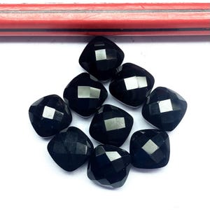 Puede incluir: Colección de nueve piedras preciosas negras facetadas y cuadradas. Las piedras están dispuestas en un patrón circular sobre una superficie blanca. Las gemas tienen una superficie pulida y reflectante.