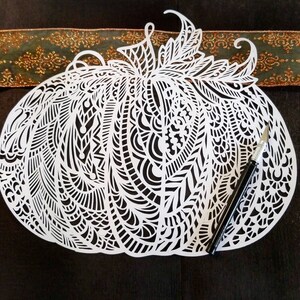 May include: A white paper pumpkin cutout with intricate, detailed designs. The pumpkin is on a dark green surface with a craft knife beside it.