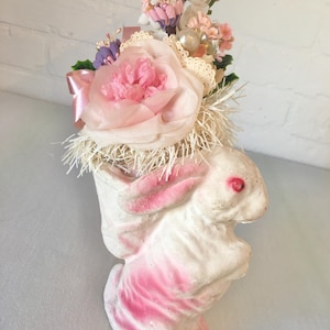 May include: A white bunny figurine with pink accents, holding a floral arrangement. The arrangement includes a large pink paper flower, lace, and small artificial flowers. A small fairy figurine sits atop the arrangement. The bunny has red eyes.