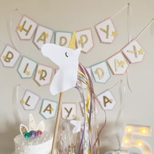Pu&ograve; includere: Una decorazione per feste di compleanno a tema unicorno con una testa di unicorno bianca su un bastoncino di legno con nastri colorati. La testa di unicorno ha un corno dorato e gli occhi chiusi. Lo sfondo include uno striscione che dice "Buon compleanno" con lettere dorate scintillanti e stelle.