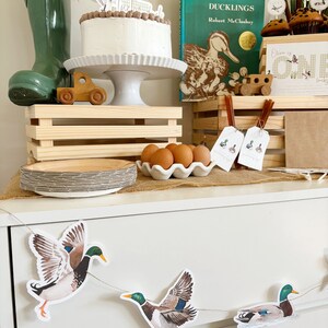 May include: A first birthday party table setting with a duck theme. The table is decorated with a cake, a book titled "Make Way for Ducklings", a wooden toy train, and a banner with the word "Duck" on it. There are also several duck-themed decorations, including a duck-shaped cake topper, a duck-shaped cookie cutter, and a duck-shaped banner. The table is set with a wooden crate, a white plate, and a white bowl filled with eggs.