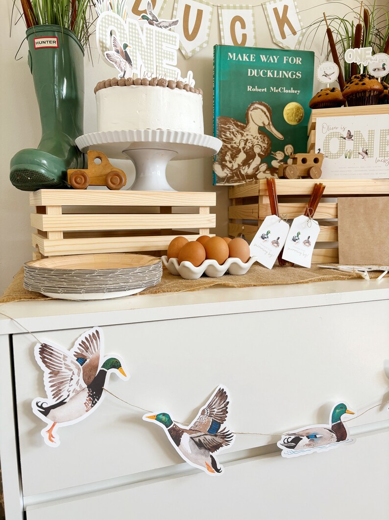 May include: A first birthday party table setting with a duck theme. The table is decorated with a cake, a book titled "Make Way for Ducklings", a wooden toy train, and a banner with the word "Duck" on it. There are also several duck-themed decorations, including a duck-shaped cake topper, a duck-shaped cookie cutter, and a duck-shaped banner. The table is set with a wooden crate, a white plate, and a white bowl filled with eggs.