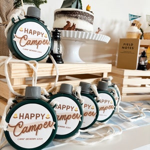 May include: A collection of green, round canteens with white rope straps. Each canteen has a white label with the words "HAPPY Camper" in brown script and the text "CAMP GRAHAM 2024". A camping-themed cake and decorations are in the background.