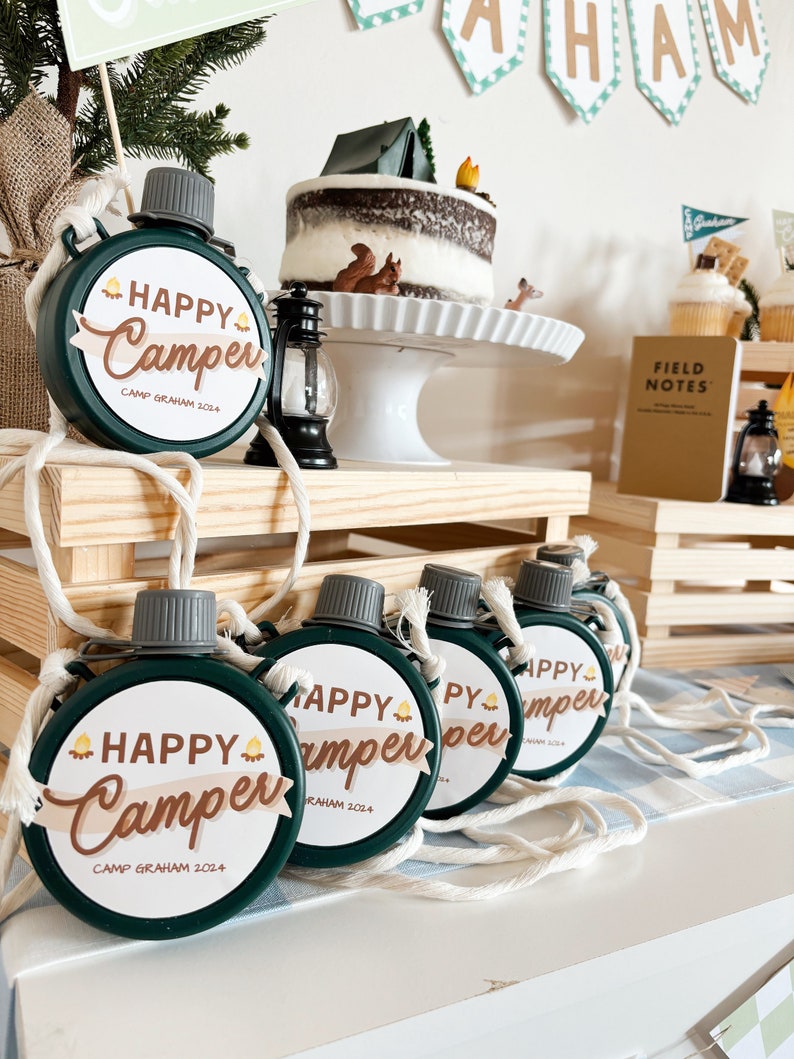 May include: A collection of green, round canteens with white rope straps. Each canteen has a white label with the words "HAPPY Camper" in brown script and the text "CAMP GRAHAM 2024". A camping-themed cake and decorations are in the background.