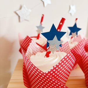 Pu&ograve; includere: Fodere per cupcake in vichy rosso e bianco con glassa bianca e un topper a stella blu e argento su una cannuccia a righe rosse e bianche.