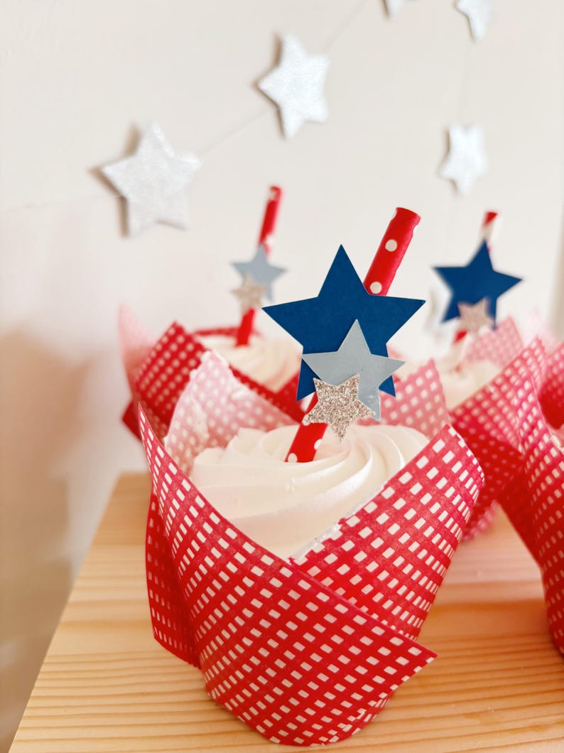 Pu&ograve; includere: Fodere per cupcake in vichy rosso e bianco con glassa bianca e un topper a stella blu e argento su una cannuccia a righe rosse e bianche.