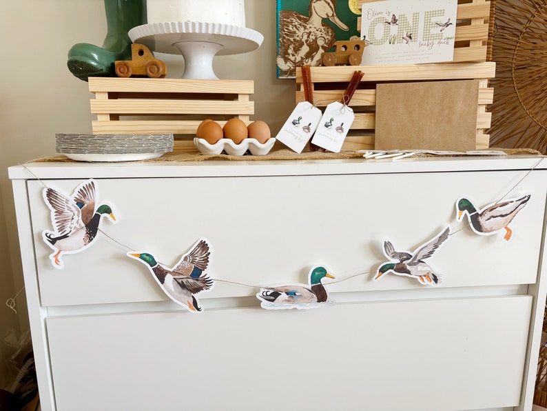 May include: A white dresser with a duck themed banner hanging above it. The banner has five ducks in flight, with green heads and brown bodies. The dresser has two drawers and is decorated with a burlap runner and a small white plate with three brown eggs.