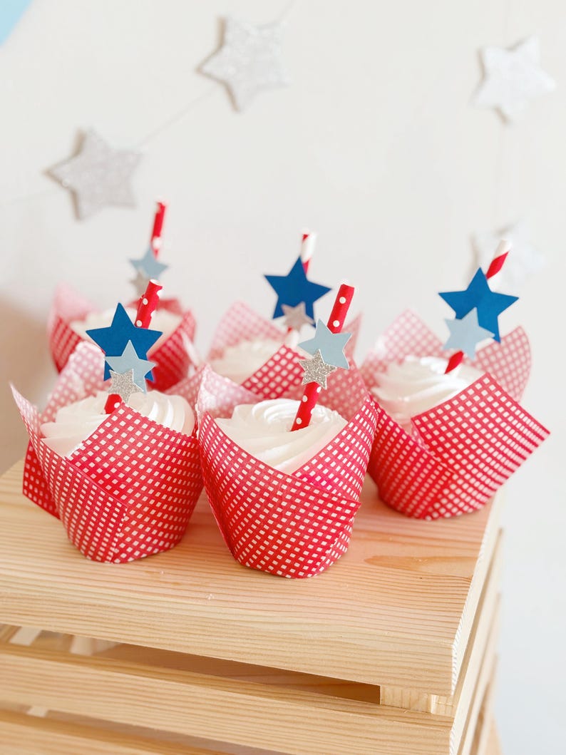 Pu&ograve; includere: Fodere per cupcake in vichy rosso e bianco con glassa bianca e decorazioni a forma di stella blu e argento. I cupcake sono disposti su un supporto a pi&ugrave; livelli in legno.