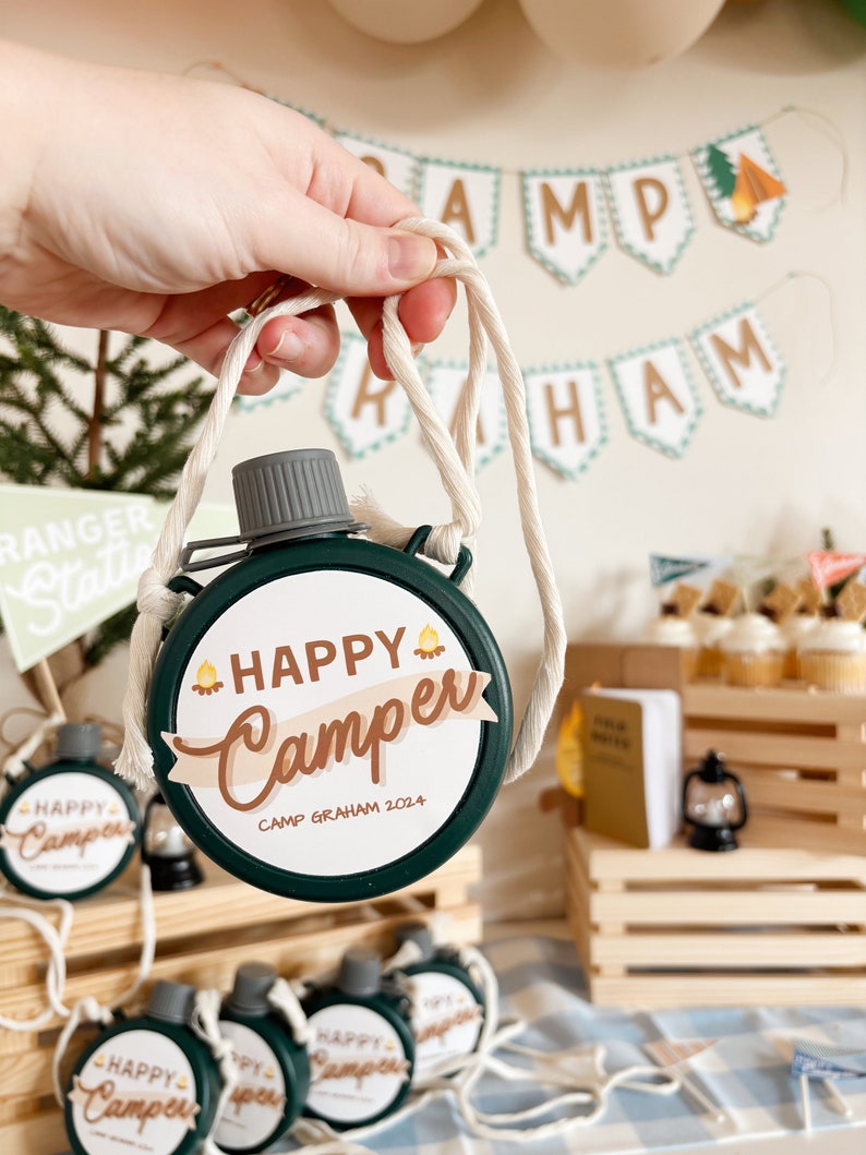 May include: A green canteen with a white label that reads "HAPPY Camper" and "CAMP GRAHAM 2024". The canteen has a beige rope strap. Other canteens and camping-themed decorations are in the background.