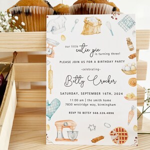 May include: A white invitation card with a watercolor illustration of baking supplies, including a mixer, rolling pin, and a pie. The text on the card reads "our little cutie pie is turning three!" and includes the name "Betty Crocker" and details for a birthday party.