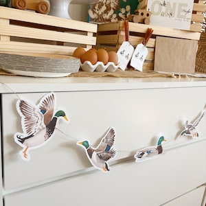 May include: A white dresser with a duck themed garland hanging on the front. The garland has five ducks, two flying and three swimming. The dresser has two drawers. The garland is made of paper and has a rustic look.