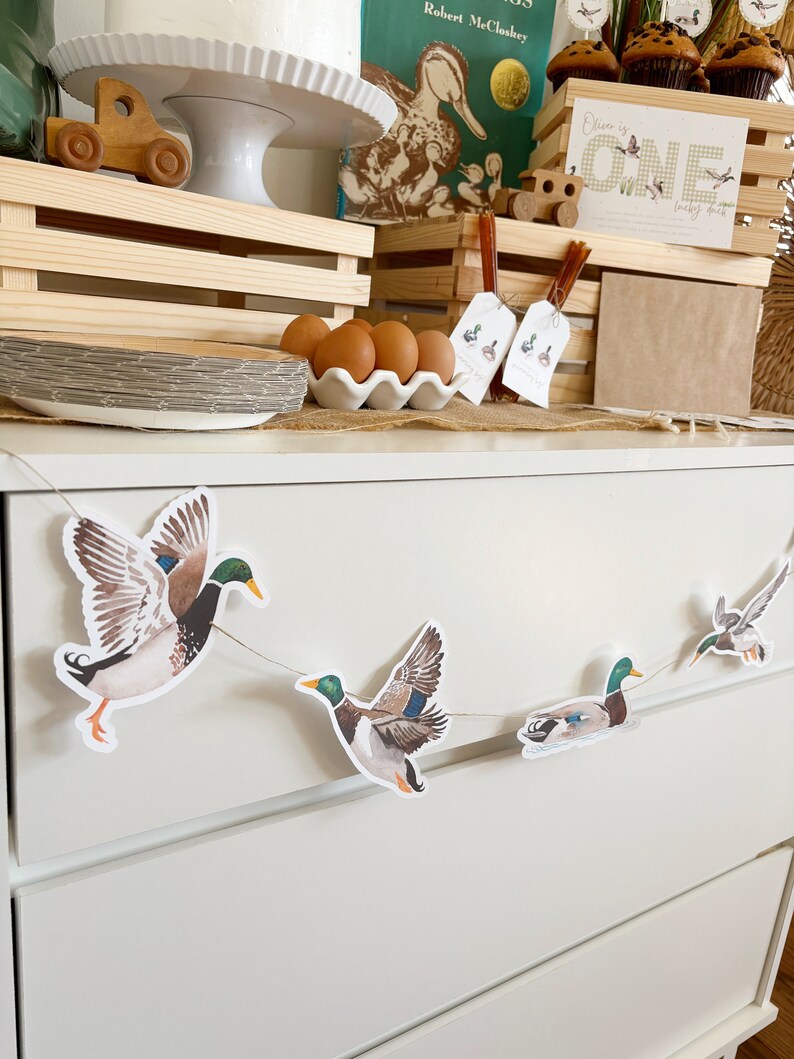 May include: A white dresser with a duck themed garland hanging on the front. The garland has five ducks, two flying and three swimming. The dresser has two drawers. The garland is made of paper and has a rustic look.