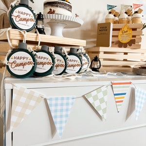 May include: A camping-themed party setup with a cake, cupcakes, and flasks. The cake is topped with a tent and trees. The flasks are green with "Happy Camper" text. A banner spells out "Graham". Cupcakes have graham cracker flags.