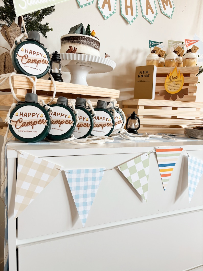 May include: A camping-themed party setup with a cake, cupcakes, and flasks. The cake is topped with a tent and trees. The flasks are green with "Happy Camper" text. A banner spells out "Graham". Cupcakes have graham cracker flags.