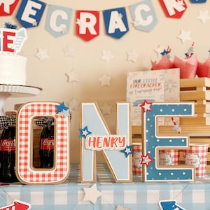 May include: A festive birthday party scene with a banner that reads "RECRACKE". The word "ONE" is spelled out with large, decorative letters. The letter "O" is red and white checkered, "N" is light blue with "HENRY" on it, and "E" is blue with white stars.