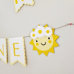 Może przedstawiać: Ż&oacute;łty papierowy baner z napisem "Summer" napisanym białymi literami. Baner jest ozdobiony ż&oacute;łtym słońcem z uśmiechniętą twarzą i białym wiankiem z stokrotek.