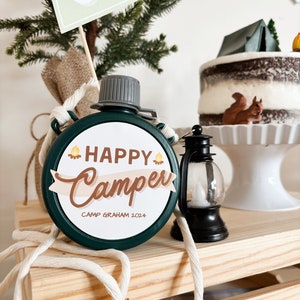 May include: A green canteen with the words "Happy Camper" and "Camp Graham 2024" printed on it. The canteen is round and has a gray cap. A small black lantern and a cake with a tent and squirrel decoration are also visible.