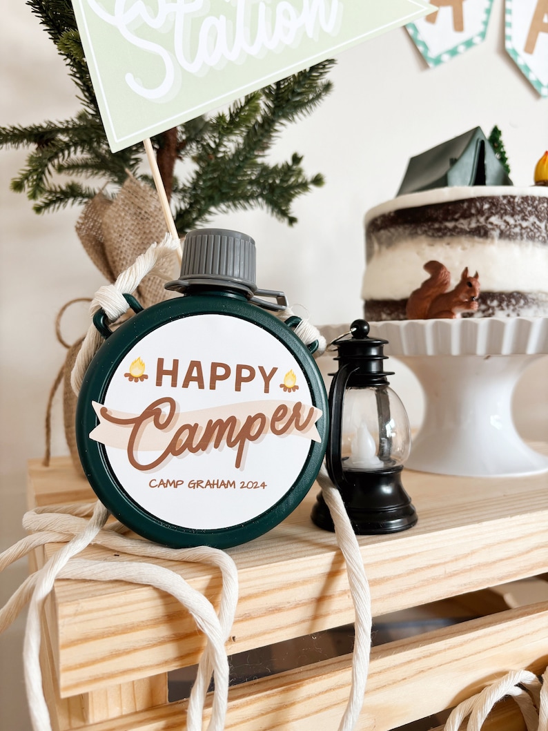 May include: A green canteen with the words "Happy Camper" and "Camp Graham 2024" printed on it. The canteen is round and has a gray cap. A small black lantern and a cake with a tent and squirrel decoration are also visible.