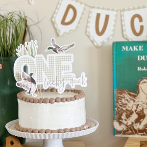 May include: A white cake with brown frosting and a 'One' cake topper with a duck and the words 'lucky duck'. The cake is on a white cake stand. There is a green book with the words 'Make Way for Ducklings' on the shelf behind the cake.