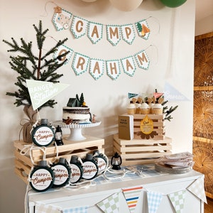 May include: A festive camping-themed party setup with a banner that reads "Camp Graham." The scene includes a cake, cupcakes, and decorative elements like wooden crates, lanterns, and pennant flags. Green and white balloons are visible in the background. Several flasks with the text "Happy Camper" are displayed.