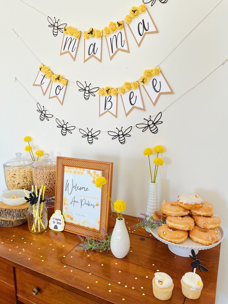 以下が含まれることがあります： 黄色と黒の蜂をテーマにしたベビーシャワーパーティーのテーブル。テーブルには「Ann Pushingのベビーシャワーへようこそ」と書かれた看板があります。テーブルにはドーナツ、カップケーキ、シリアルが置かれています。