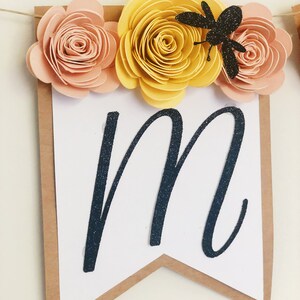以下が含まれることがあります： 黒いグリッターで「M」の文字が書かれた白いバナー。黄色と桃色の紙の花と黒いグリッターの蜂で飾られています。