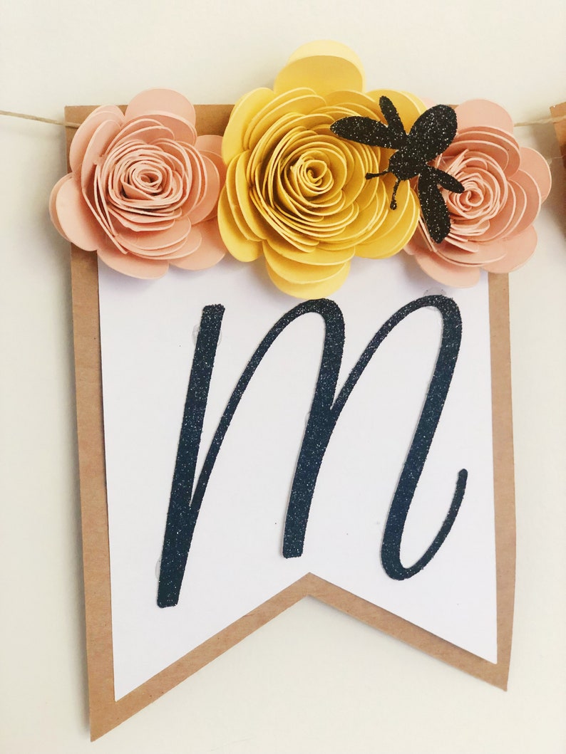 以下が含まれることがあります： 黒いグリッターで「M」の文字が書かれた白いバナー。黄色と桃色の紙の花と黒いグリッターの蜂で飾られています。
