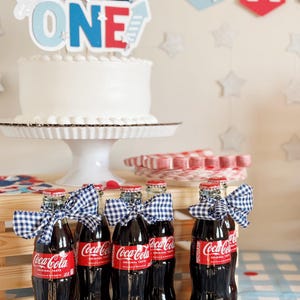 以下が含まれることがあります： 青、赤、白の「ONE」の飾り付けがされた白いケーキ。ケーキは白いケーキスタンドの上に置かれています。青と白のギンガムチェックのリボンが付いたコカ・コーラのガラス瓶が6本あります。瓶は木製の箱と青と白のチェックのテーブルクロスの上に置かれています。