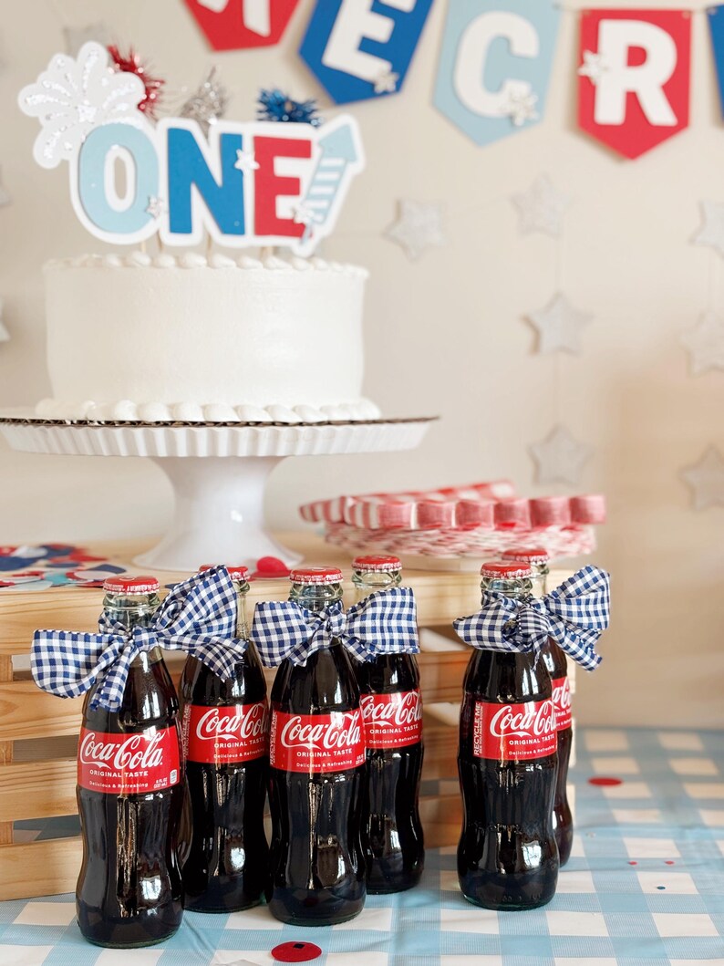 以下が含まれることがあります： 青、赤、白の「ONE」の飾り付けがされた白いケーキ。ケーキは白いケーキスタンドの上に置かれています。青と白のギンガムチェックのリボンが付いたコカ・コーラのガラス瓶が6本あります。瓶は木製の箱と青と白のチェックのテーブルクロスの上に置かれています。