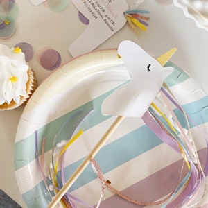 Pu&ograve; includere: Un favore per feste di unicorno bianco con un corno dorato e un bastone di legno. L'unicorno &egrave; attaccato a un piatto di carta a strisce colorate con nastri rosa, blu e gialli.