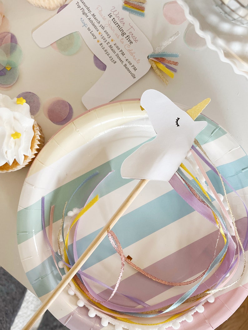 Pu&ograve; includere: Un favore per feste di unicorno bianco con un corno dorato e un bastone di legno. L'unicorno &egrave; attaccato a un piatto di carta a strisce colorate con nastri rosa, blu e gialli.