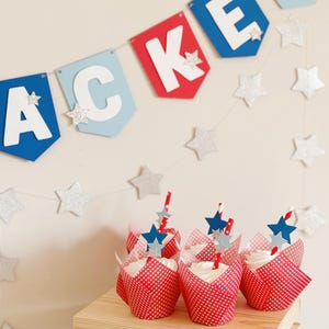 Pu&ograve; includere: Fodere per cupcake rosso, bianco e blu con glassa bianca e decorazioni a forma di stella blu. I cupcake sono disposti su una cassa di legno con uno striscione rosso, bianco e blu che dice "Jacker" appeso sopra.