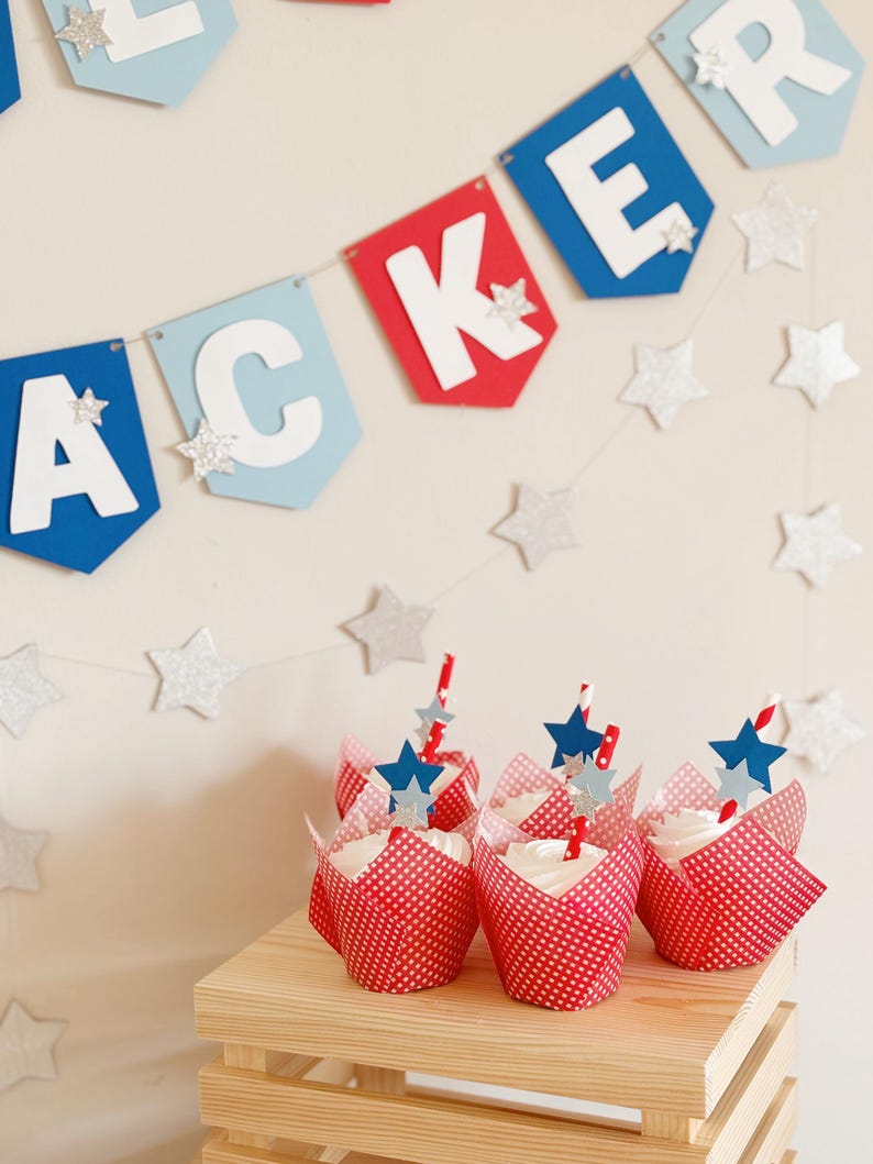 Pu&ograve; includere: Fodere per cupcake rosso, bianco e blu con glassa bianca e decorazioni a forma di stella blu. I cupcake sono disposti su una cassa di legno con uno striscione rosso, bianco e blu che dice "Jacker" appeso sopra.