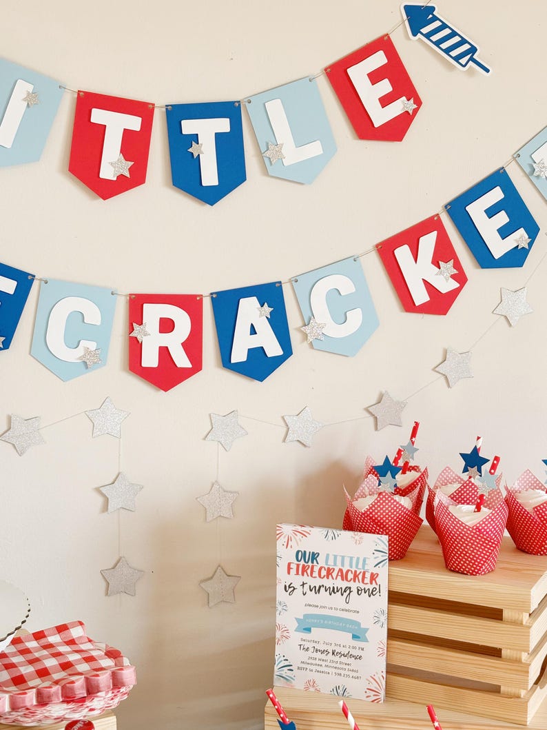 Pu&ograve; includere: Uno striscione rosso, bianco e blu con le parole "Little Firecracker" e stelle argentate scintillanti. Un invito a una festa con il testo "Our Little Firecracker is turning one!" &egrave; su una cassa di legno con pirottini per cupcake a pois rossi e bianchi.