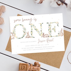 May include: A white invitation card with a floral wreath spelling out "ONE" with a bunny rabbit in each letter. The text "some bunny is" is above the wreath and the text "Jessica Rabbit" is below the wreath. The invitation details a first birthday party with the date, time, location, and RSVP information.