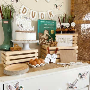 May include: A first birthday party table setting with a duck theme. The table is decorated with a banner that says "Lucky Duck", a cake with a duck on top, and cupcakes with duck toppers. There is also a book titled "Make Way for Ducklings" by Robert McCloskey.