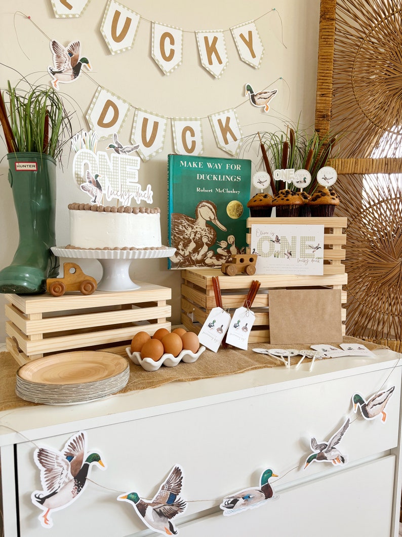 May include: A first birthday party table setting with a duck theme. The table is decorated with a banner that says "Lucky Duck", a cake with a duck on top, and cupcakes with duck toppers. There is also a book titled "Make Way for Ducklings" by Robert McCloskey.