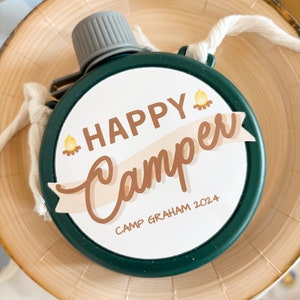May include: A green, round canteen with a gray cap and a white cord. The canteen has a white circular label with the words "HAPPY Camper" and "Camp Graham 2024" in brown lettering, with a campfire graphic. The canteen is on a wooden plate.