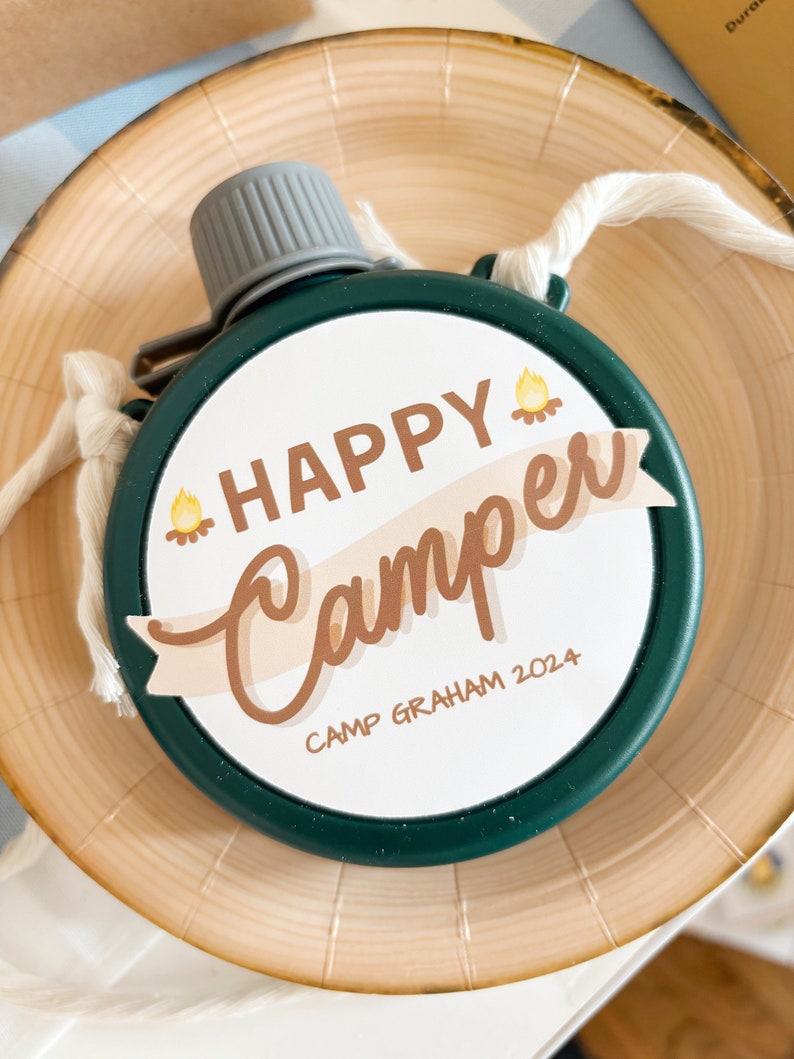 May include: A green, round canteen with a gray cap and a white cord. The canteen has a white circular label with the words "HAPPY Camper" and "Camp Graham 2024" in brown lettering, with a campfire graphic. The canteen is on a wooden plate.