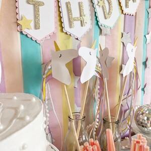 Pu&ograve; includere: Un set di decorazioni per una festa di compleanno a tema unicorno con uno striscione che recita "Buon compleanno" in lettere dorate glitterate. Lo striscione &egrave; appeso su uno sfondo a strisce pastello. Ci sono anche ritagli di teste di unicorno e bacchette con nastri colorati.