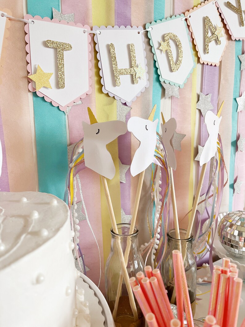 Pu&ograve; includere: Un set di decorazioni per una festa di compleanno a tema unicorno con uno striscione che recita "Buon compleanno" in lettere dorate glitterate. Lo striscione &egrave; appeso su uno sfondo a strisce pastello. Ci sono anche ritagli di teste di unicorno e bacchette con nastri colorati.