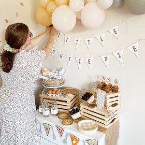 May include: A baby shower party setup with a white banner that reads "Little Cutie Pie". The banner is decorated with brown letters and a scalloped edge. There are balloons in shades of beige and gray, and a table with a variety of food and decorations. The table is decorated with wooden crates, a checkered tablecloth, and a variety of food items, including pies, cupcakes, and cherries.