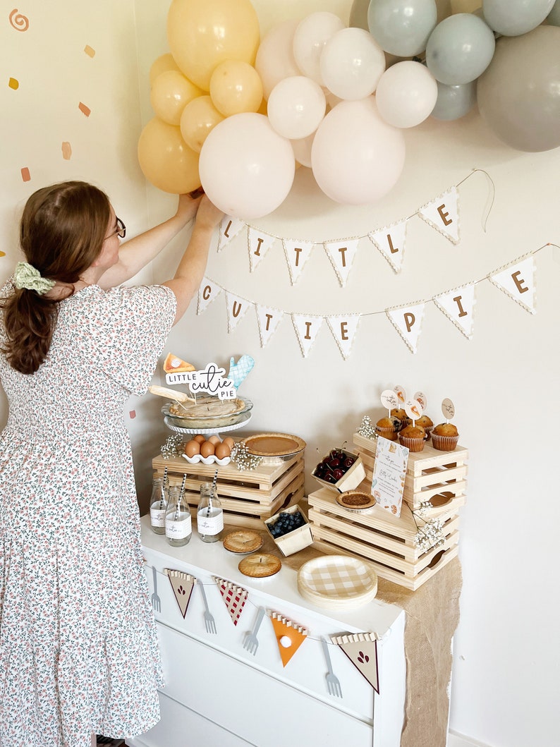 May include: A baby shower party setup with a white banner that reads "Little Cutie Pie". The banner is decorated with brown letters and a scalloped edge. There are balloons in shades of beige and gray, and a table with a variety of food and decorations. The table is decorated with wooden crates, a checkered tablecloth, and a variety of food items, including pies, cupcakes, and cherries.