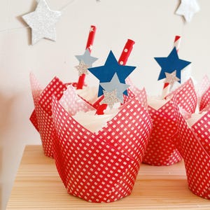 Pu&ograve; includere: Fodere per cupcake a quadri rossi e bianchi con glassa bianca e decorazioni a forma di stella blu e argento. I cupcake sono sormontati da cannucce a righe rosse e bianche.
