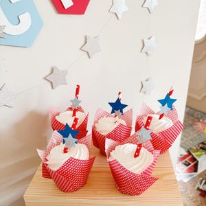 Pu&ograve; includere: Cinque pirottini per cupcake a pois rossi e bianchi con glassa bianca e decorazioni a forma di stella blu e argento. I cupcake sono disposti su un supporto a pi&ugrave; livelli in legno.